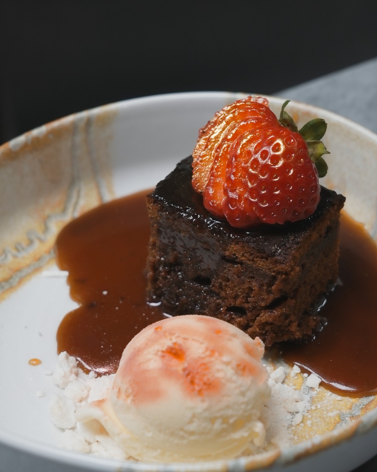 Sticky toffee pudding crowned with a fanned strawberry, toffee sauce pooling around it and vanilla ice cream on meringue, on a dark table
